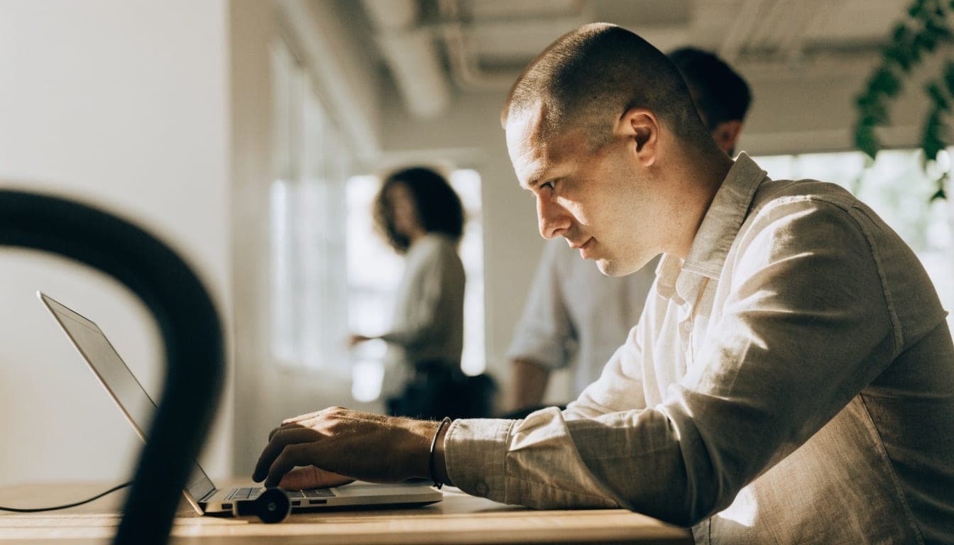 Man werkt geconcentreerd op een laptop in een kantoor met collega's op de achtergrond