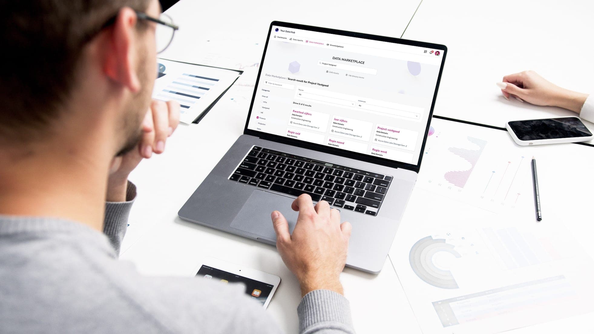 Twee collega’s aan een tafel; op het laptopscherm staat de Data Marketplace met een zoekopdracht naar een project en de gevonden datasets per regio.