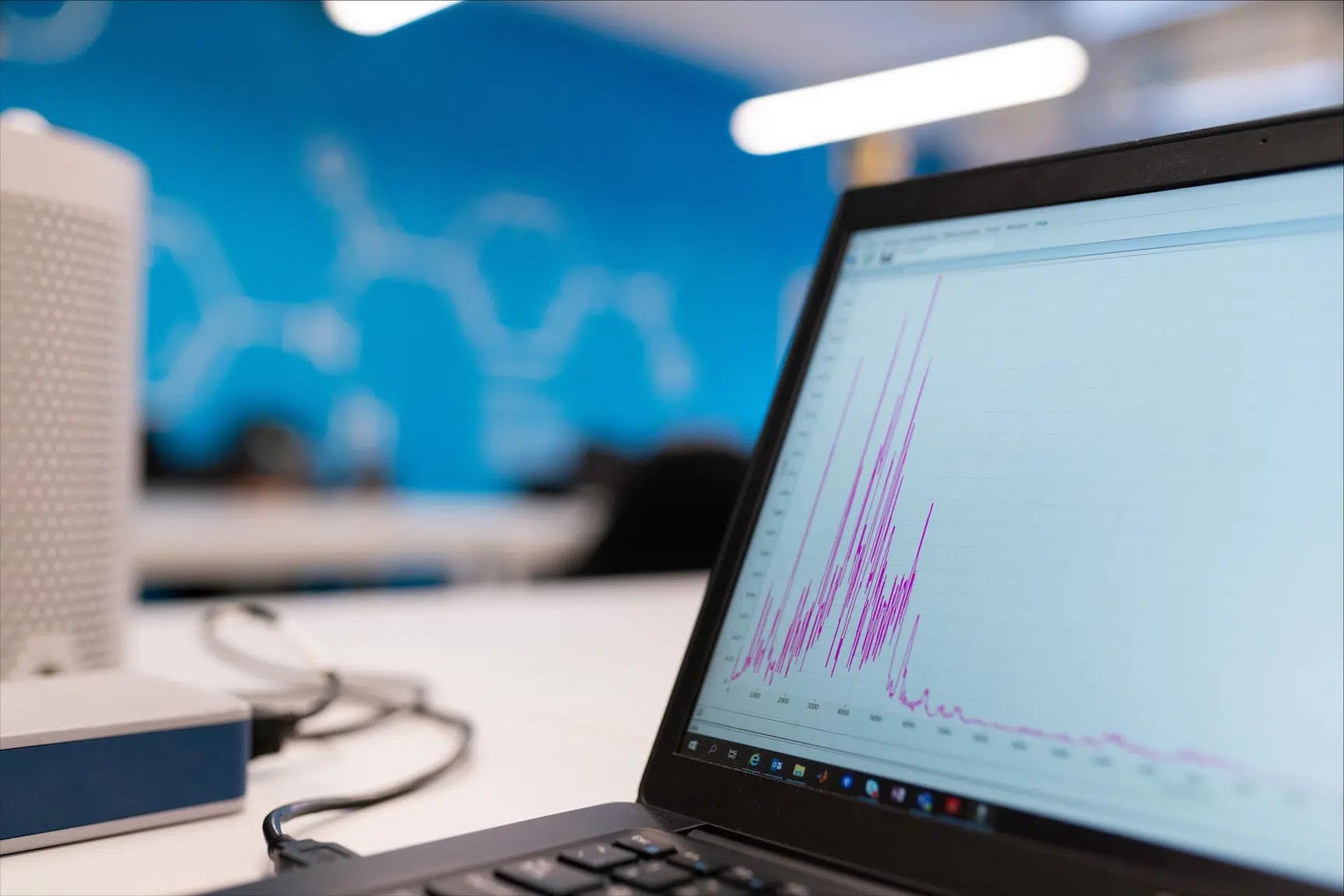Een laptop op een bureau toont een grafiek met scherpe pieken; op de achtergrond een blauwe wand met een datavisualisatie.