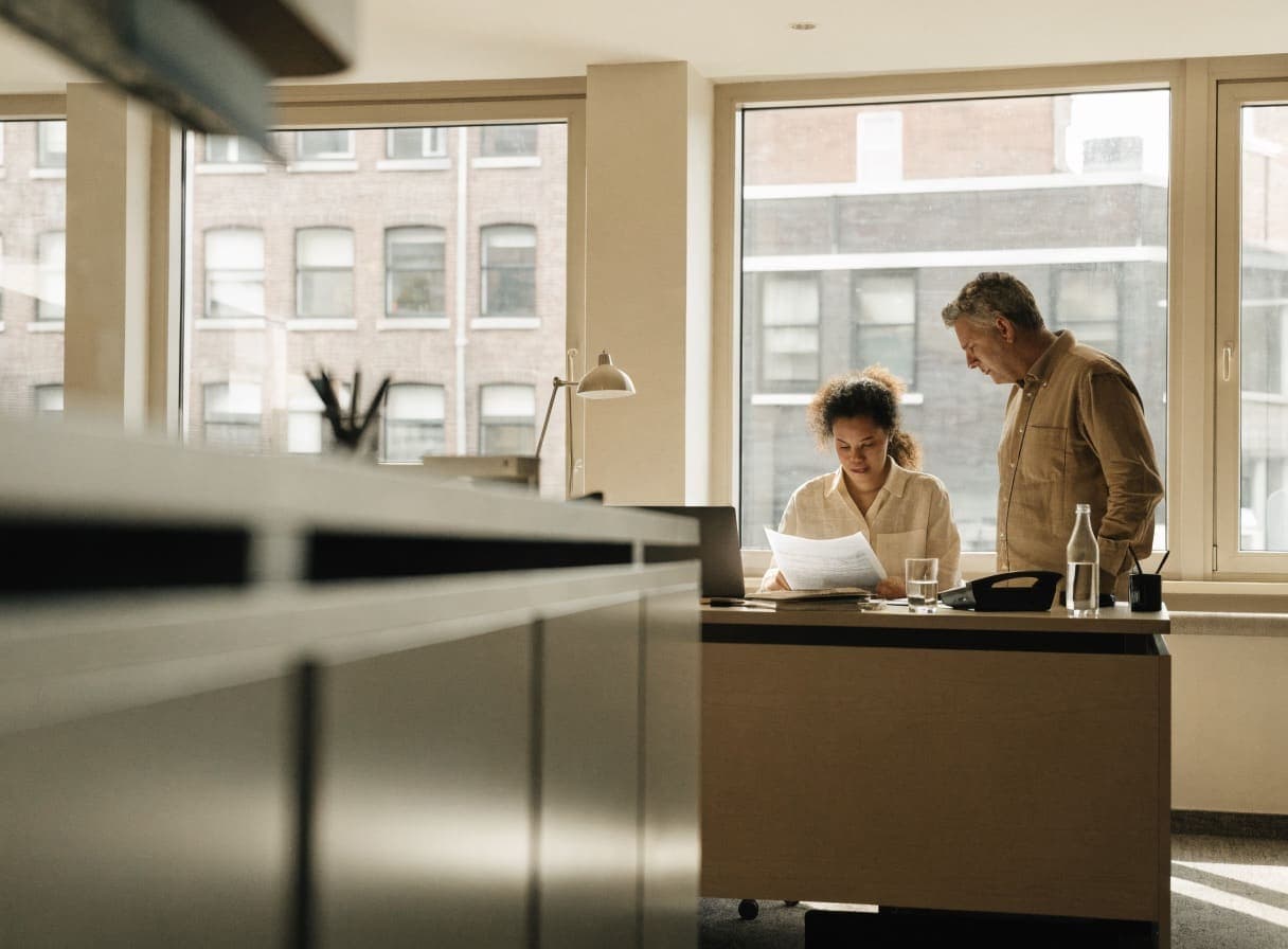 Medewerker leest een document aan haar bureau met een laptop, terwijl een collega meekijkt